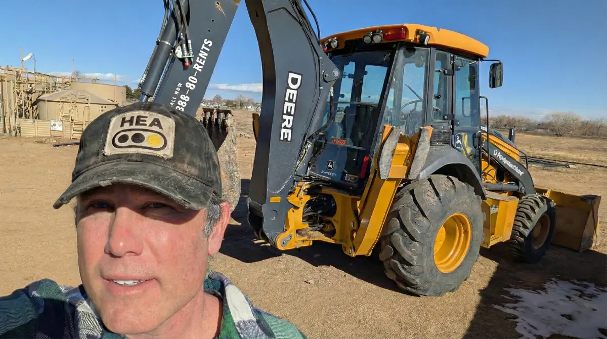 A CMEA (Certified Machinery and Equipment Appraiser) from Heavy Equipment Appraisal (HEA) performing an on-site, physical inspection of a John Deere backhoe loader. The appraiser, wearing a branded HEA hat, is on a dirt construction site to physically verify the machinery’s condition, serial number, and engine hours for a USPAP-compliant valuation report.