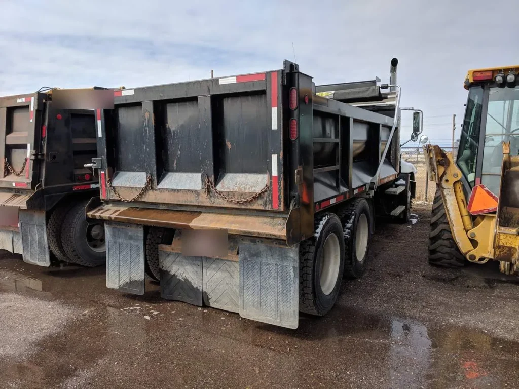 Rear three-quarter view of a tandem-axle dump truck body and frame beside a second dump truck and a backhoe
