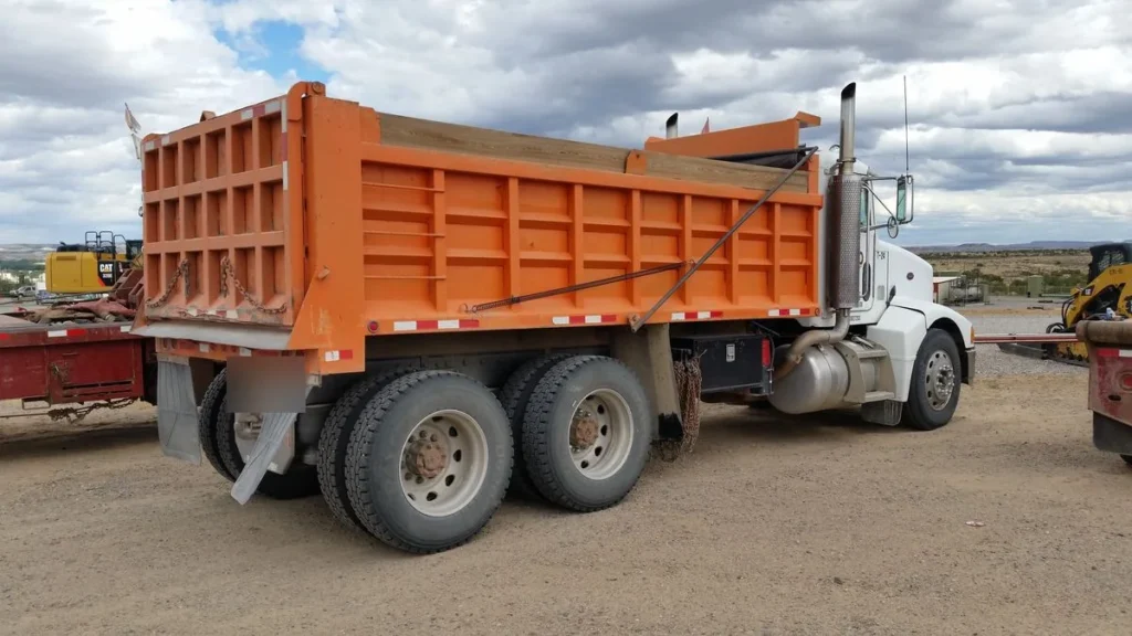 Tandem-axle dump truck with an orange steel dump body, rear three-quarter view in a New Mexico yard